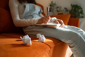 Young woman sits on an orange sofa, diligently writing in a notebook while several crumpled pieces of paper lie beside her, suggesting a creative struggle or writer's block